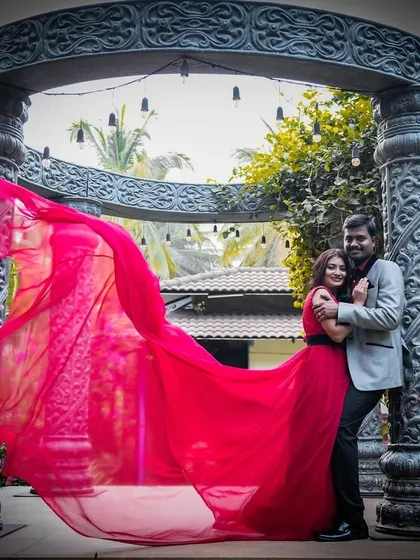 A romantic pre-wedding embrace under an ornate archway. The flowing red trail of the gown adds a touch of drama and passion.