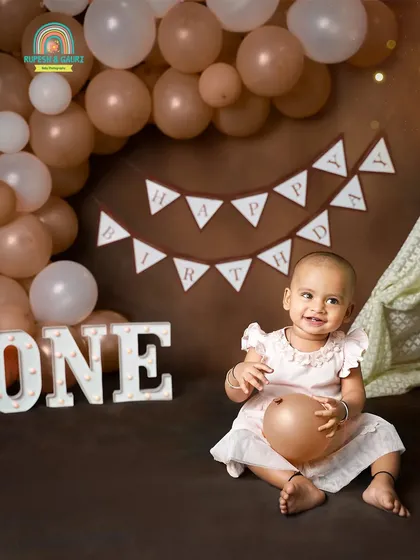 A happy one-year-old in our studio, ready for her cake smash. The rustic brown and balloon setup creates a warm and celebratory atmosphere.
