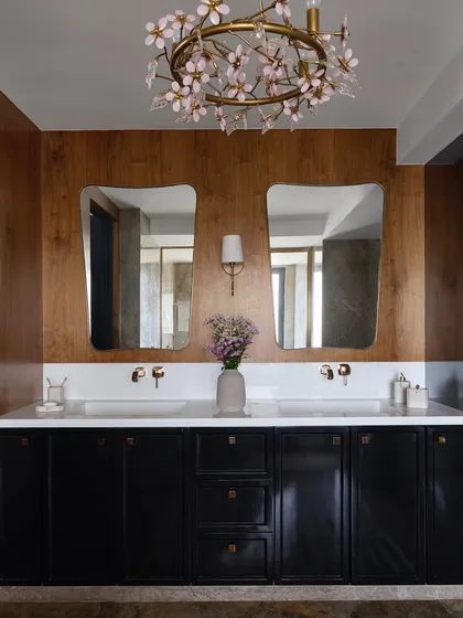 This bathroom is all about contrast, with a rich brown vanity and a black cabinet set against a veneered wood wall. The dual sinks and a delicate floral-inspired chandelier soften the space.