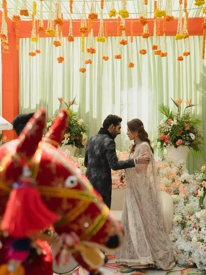 A candid moment between the couple during their Mehendi, framed by a traditional red horse figurine. The decor features a canopy of hanging marigolds and lush floral arrangements.