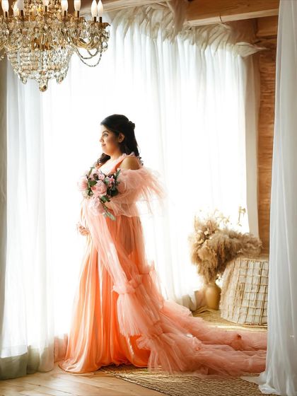 A full-length portrait showcasing the dramatic train of a flowing peach gown against a brightly lit window.