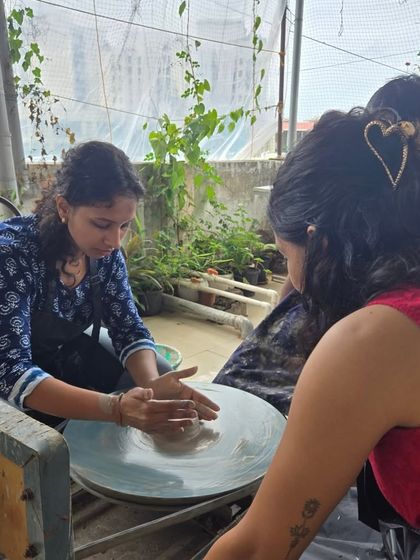 A participant learns the basics of wheel throwing, her hands gently guiding the clay. The lush green plants in the background highlight our open, airy studio space.