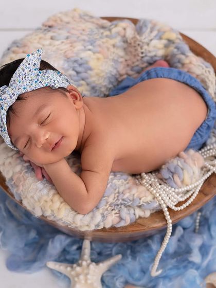 A smiling newborn with a cute blue bow headband sleeps in a wooden bowl, with pearls and starfish props hinting at a subtle beach theme.