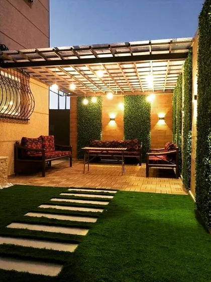 A narrow yard transformed with a pergola, artificial turf, and clever lighting. The back wall combines wood paneling and vertical gardens, illuminated by string lights and wall sconces to make the space feel larger.