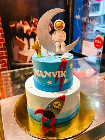 A two-tier astronaut cake for Manvik's first birthday, with a rocket ship and a cute astronaut sitting on the moon.