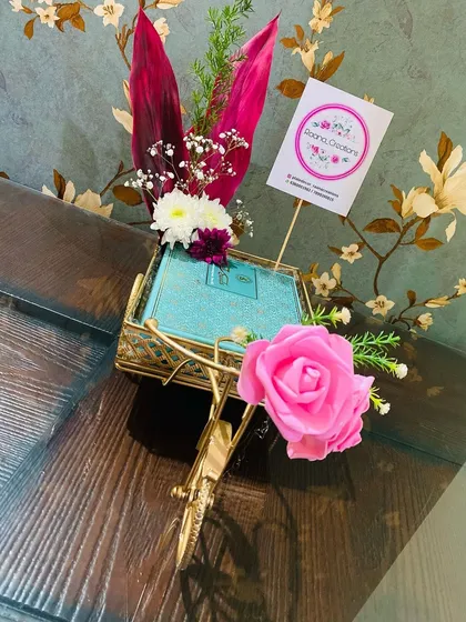 We love finding creative ways to present your return gifts. Here, a gift box is presented in a charming, gold-finished miniature cycle rickshaw, decorated with a pink rose for a whimsical touch.