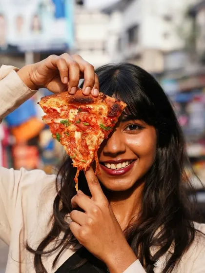 A fun and relatable lifestyle shot of a woman smiling while holding up a slice of cheesy pizza, covering one eye. This image captures the pure joy of eating and is perfect for social media content that connects with customers on a personal level.