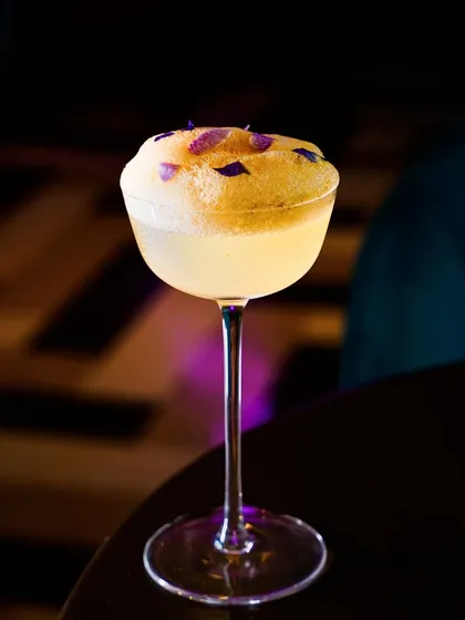 A classic sour cocktail with a perfect foam top, garnished with edible flowers, set against a dark background with purple and blue neon lights. This image captures a sophisticated, modern bar atmosphere.