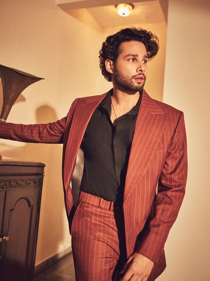 Siddhant Chaturvedi posing next to a gramophone, which complements the vintage feel of his rust-colored pinstripe suit. It’s about creating a whole mood, not just an outfit.