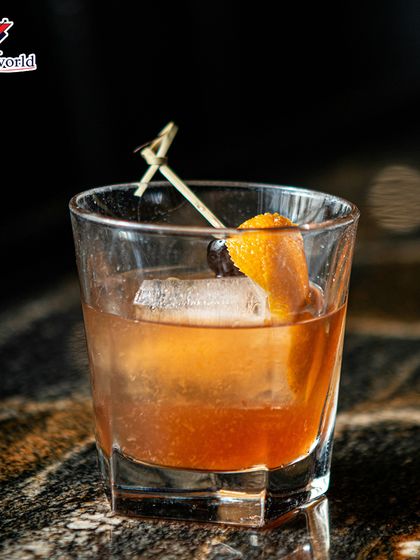 An Old Fashioned served over a large ice cube with a classic orange peel and cherry garnish. This is a timeless drink, expertly crafted for the discerning palate.