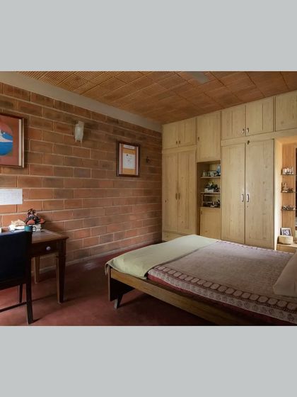 A bedroom featuring an exposed brick wall, red oxide flooring, and simple wooden furniture, creating a warm, earthy, and restful space.