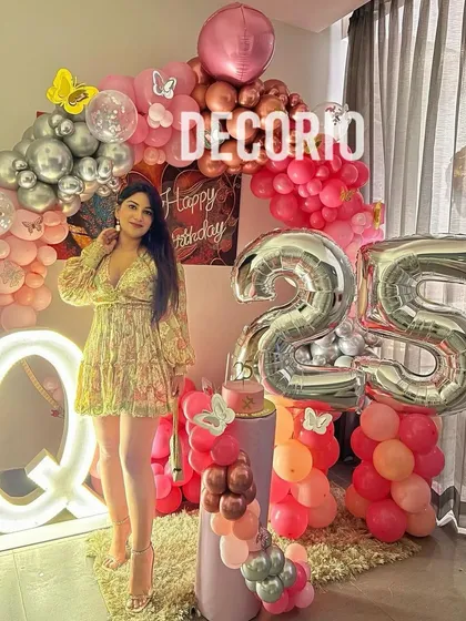 The birthday girl posing with her stunning 25th birthday decor. The setup includes large silver foil '25' balloons, a lit 'Q' marquee letter, and an elaborate pink and silver balloon arch.