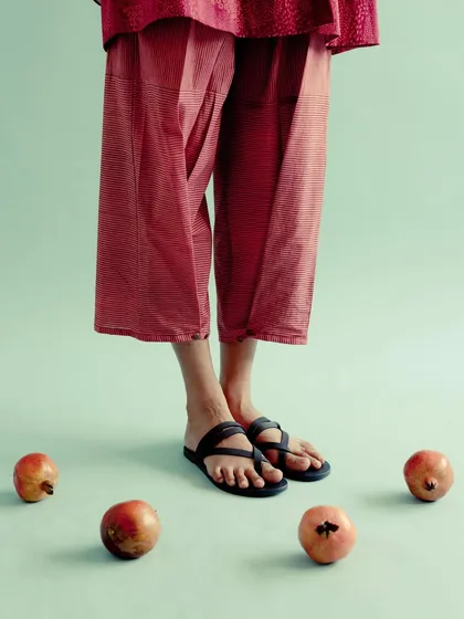 A minimalist detail shot for Swatti Kapoor. The focus is on the feet, showcasing the simple sandals and cropped trousers against a clean background with scattered pomegranates.