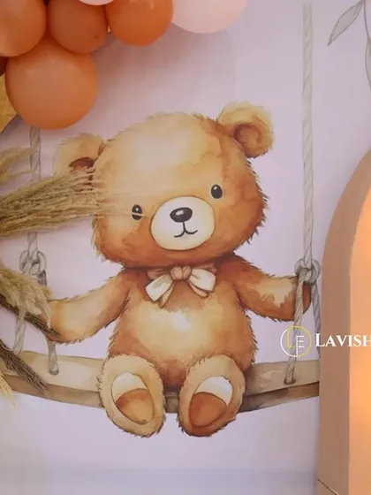 A close-up of a custom printed backdrop featuring a cute teddy bear on a swing, complemented by dried palm leaves and marquee lights.