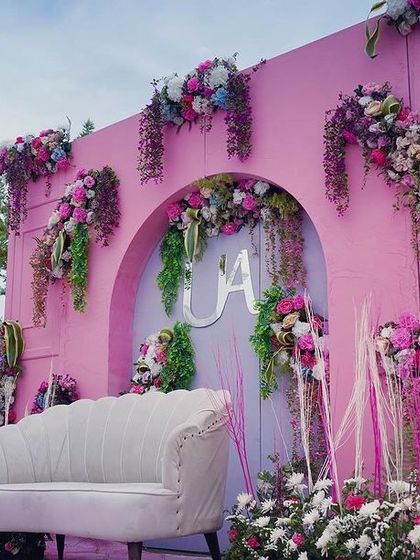 The main stage for a pastel carnival Mehendi, set against a stunning mountain view. The backdrop is a vibrant pink wall adorned with cascading florals and a custom monogram for the couple.