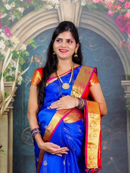 A beautiful portrait of a mother-to-be in a traditional blue and red saree. The floral studio backdrop adds a touch of softness and celebration to the image.