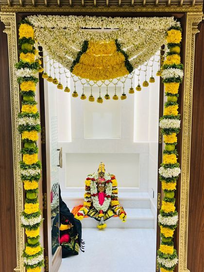A full view of a pooja room entrance decorated for a housewarming. The deity is visible through the door, framed perfectly by the auspicious and beautiful floral 'thomala'.