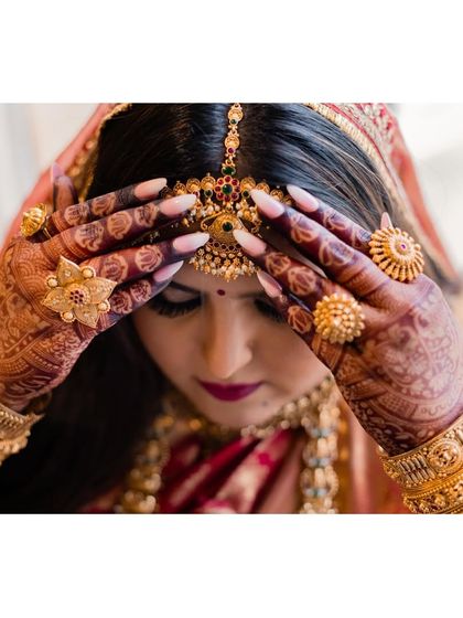 A close-up of the bride's final touches. Her hands, adorned with henna and gold rings, adjust the intricate temple jewelry, a key element of the South Indian bridal aesthetic.