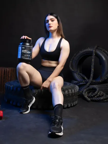 A full-body shot of the female model seated among gym props, holding the whey protein. This composition provides a complete lifestyle context, showing how the product fits into the world of a dedicated fitness enthusiast.
