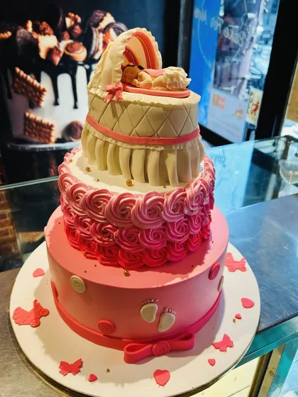 A three-tier pink baby shower cake with a quilted bassinet topper holding a sleeping baby.