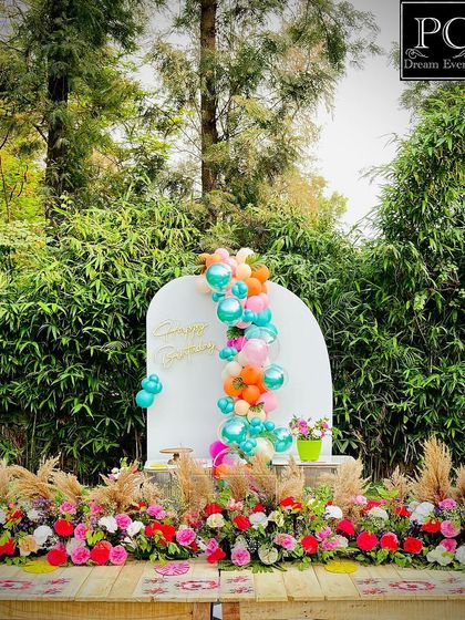 The main decor for an outdoor birthday party, featuring a long rustic table decorated with flowers and a modern arched backdrop with a colorful balloon garland.