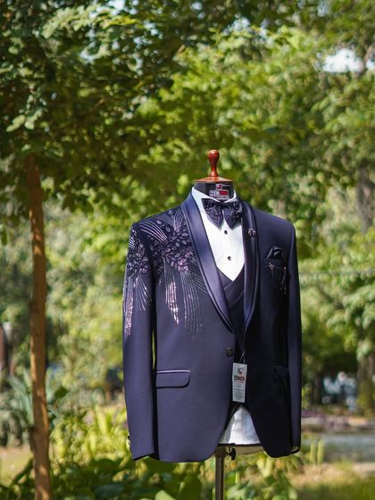 A full shot of a navy blue three-piece tuxedo displayed outdoors. The unique silver and purple feather-like embroidery on the shoulder and lapel makes this a standout piece for any formal event.