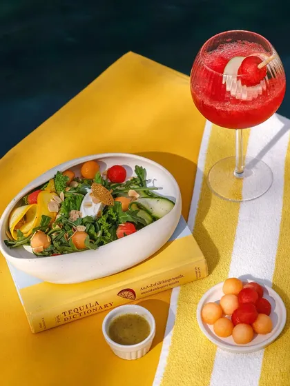 A refreshing poolside scene with a colorful salad, melon balls, and a vibrant red cocktail.