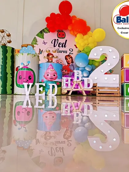 A complete Cocomelon-themed second birthday setup with a reflective floor, creating a stunning visual effect. The decor includes a rainbow balloon arch, character-printed plinths, and a large light-up number '2'.