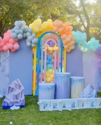 A large-scale outdoor Frozen-themed 7th birthday party. The setup includes character cutouts, ice-themed props, and a pastel rainbow balloon arch.