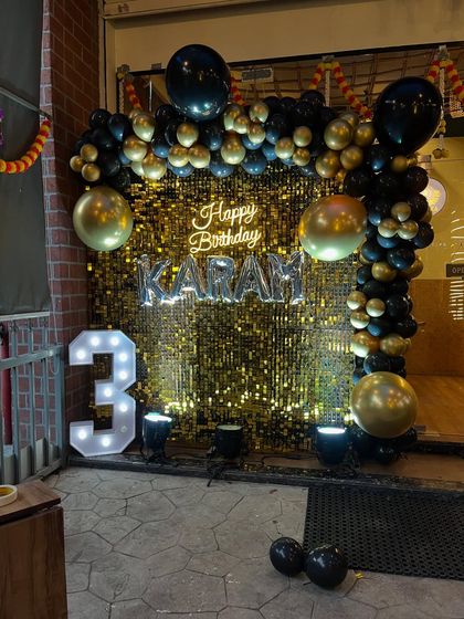 A stylish black and gold themed third birthday party. The combination of a gold shimmer wall, a neon "Happy Birthday" sign, and a black balloon arch gives it a premium feel.