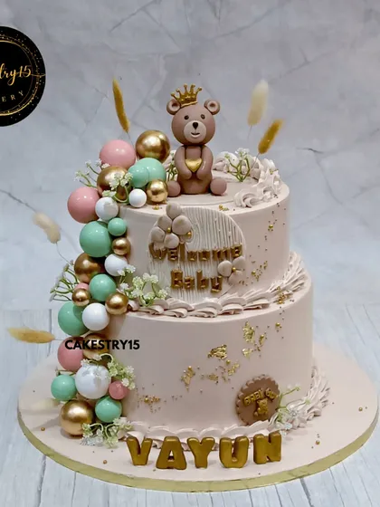 A royal teddy welcome cake for a new baby. This 5kg choco-vanilla cake is elegantly designed with a crowned teddy bear, a cascade of decorative balls, and gold leaf accents.