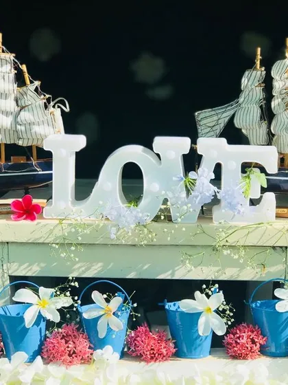 Thematic decor elements that bring the Santorini vibe to life. A light-up 'LOVE' sign is surrounded by miniature sailboats and small blue buckets with flowers, creating a romantic, nautical tablescape.