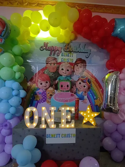 A colorful and fun Cocomelon themed first birthday setup for an at-home celebration. This decor includes a character-filled backdrop, a rainbow balloon arch, and light-up 'ONE' marquee letters.