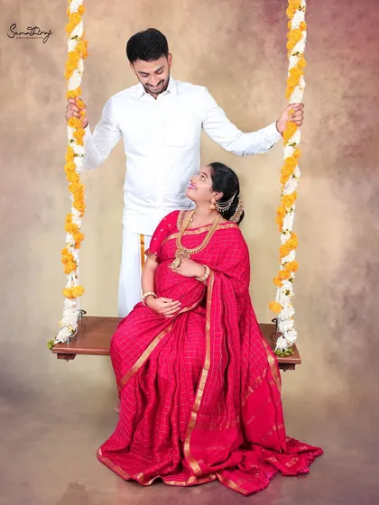 A loving gaze between a couple during their traditional maternity shoot. The beautiful red saree and the flower-adorned swing create a romantic and timeless setting.