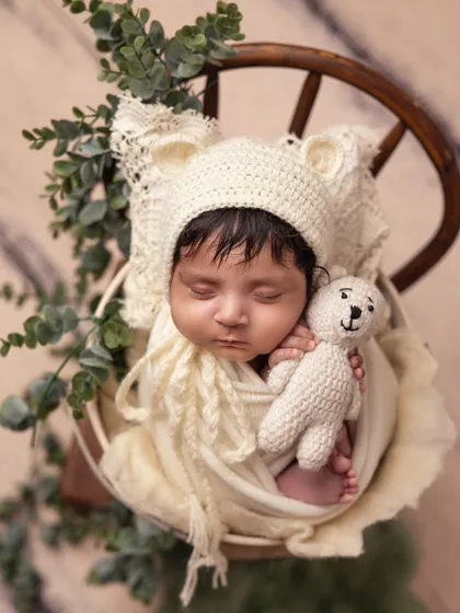 A close-up shot of a baby in a cream-colored wrap and a bear bonnet, holding a tiny knitted teddy bear. The neutral tones create a soft and timeless feel.