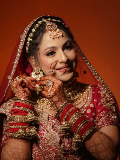 A candid moment of a bride getting ready. This image captures the rich detail of her red and gold embroidered lehenga, traditional chooda, and the warm tones of her makeup.