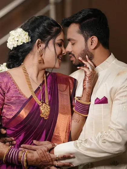 A beautiful, intimate moment between an engaged couple. Her makeup is classic and elegant, with a focus on defined eyes and a glowing complexion that complements her purple silk saree.