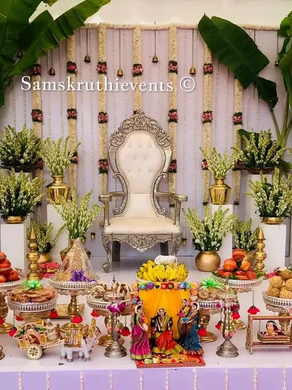 An elegant baby shower stage with a white backdrop and hanging tuberose garlands. The setup is flanked by large banana plants and features a beautiful display of offerings on a table decorated with cow figures.