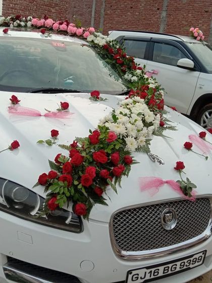 A beautiful floral design on a white Jaguar. The arrangement flows diagonally across the car, using red and white roses with delicate pink ribbons for an elegant touch.