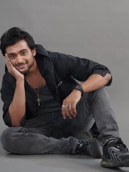 A warm and friendly seated portrait against a grey studio background. The relaxed pose and smile make this a very approachable headshot for an actor.