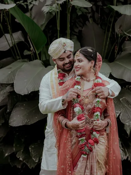 The groom helps the bride with her garland in this candid wedding portrait. The natural, leafy background contrasts beautifully with their traditional wedding attire.