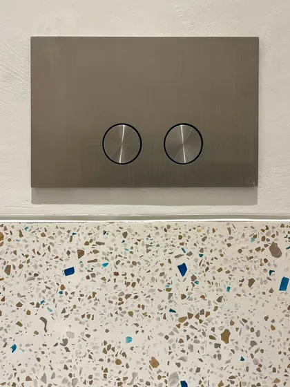 A minimalist flush plate is set against a colorful terrazzo wall. This juxtaposition of a sleek, modern fitting with a playful, textured material is a deliberate design choice to create visual interest.