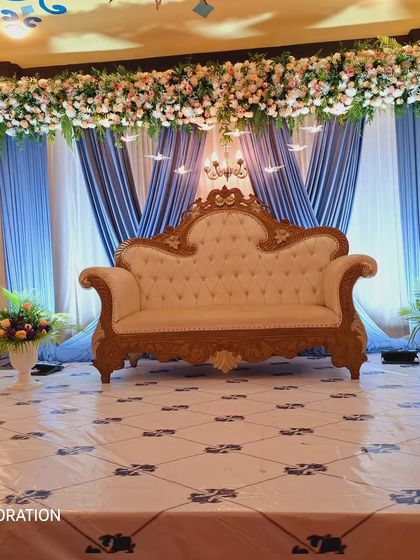 A full view of a reception stage setup in a convention hall. The design uses a striking combination of blue and white drapes, a dense overhead floral arrangement, and a classic white and gold sofa, creating a grand and elegant look.