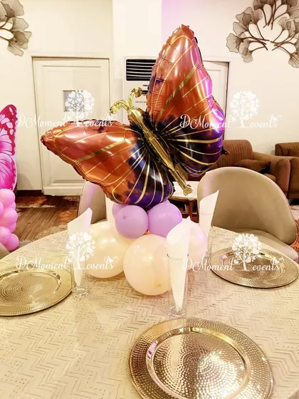 A simple yet elegant table centerpiece for a butterfly-themed party. A large, metallic butterfly foil balloon sits atop a small cluster of pastel balloons, adding a thematic touch to each table.