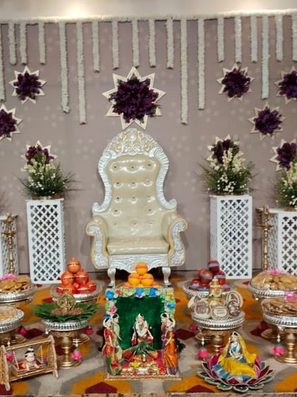 A complete and elegant baby shower setup with a beautiful white throne. The backdrop features hanging jasmine garlands and purple floral stars, complemented by traditional dolls and brass lamps.