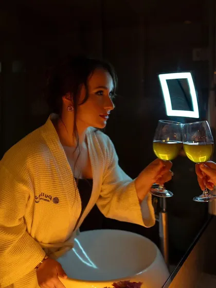 A low-light, intimate portrait of a woman in a bathrobe. The warm, candle-like lighting and the act of clinking glasses create a romantic and cozy atmosphere.