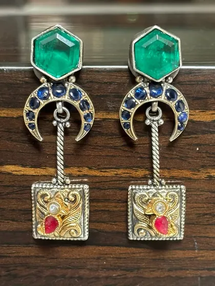 Uniquely crafted silver fusion earrings. This pair combines geometric shapes with traditional motifs, featuring emerald-green and sapphire-blue stones in a dual-tone setting of silver and gold.