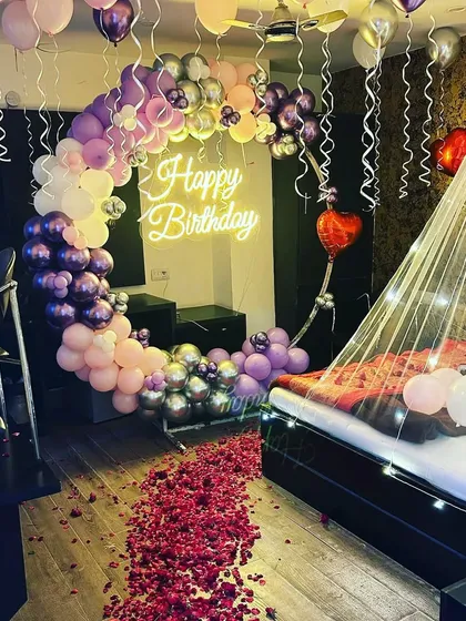 For a truly romantic birthday surprise, I transformed this room with a dreamy setup. It features a beautiful crescent moon balloon arch in shades of purple and pink, a 'Happy Birthday' neon sign, and a delicate canopy over the bed with fairy lights. The floor covered in rose petals completes the magical look.