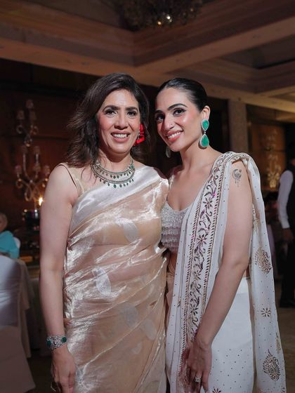 Posing with my mother at a wedding. I’m wearing a white block-printed saree with bold green earrings, while she looks elegant in a tissue saree.