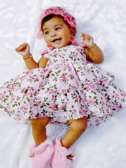 An adorable baby girl smiling in her pink floral tiered dress with a matching bonnet. My collection includes sweet styles for even the littlest fashionistas.